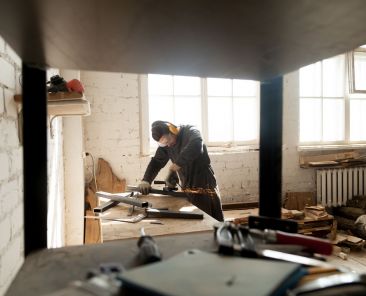 Trained young workman grinding metal parts of construction on table in workshop interior with tools equipment. Manual worker wearing protective clothes working with angle grinder in manufactory