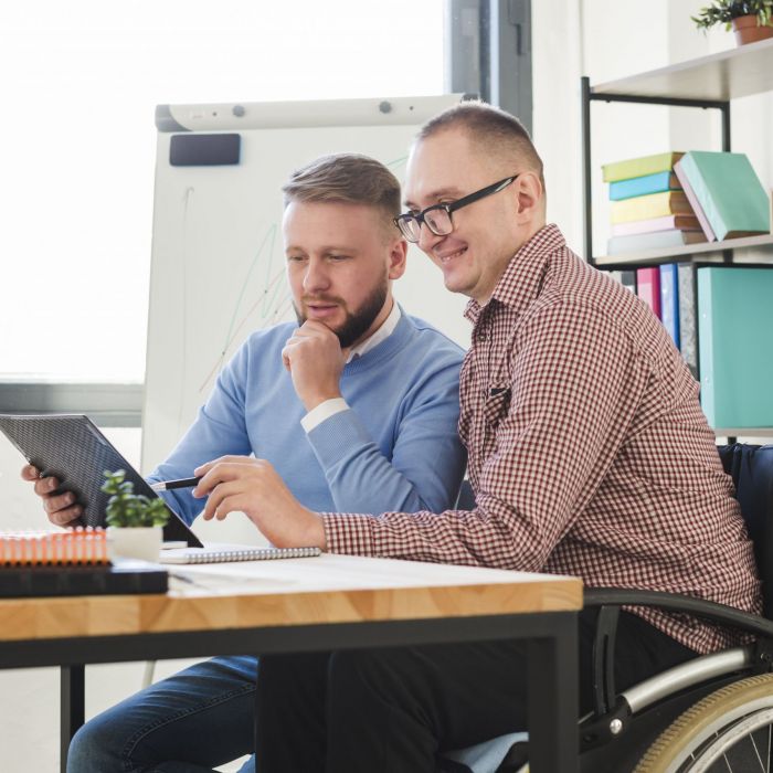 zwei Männer sitzen an einem Schreibtisch. Einer davon im Rollstuhl.